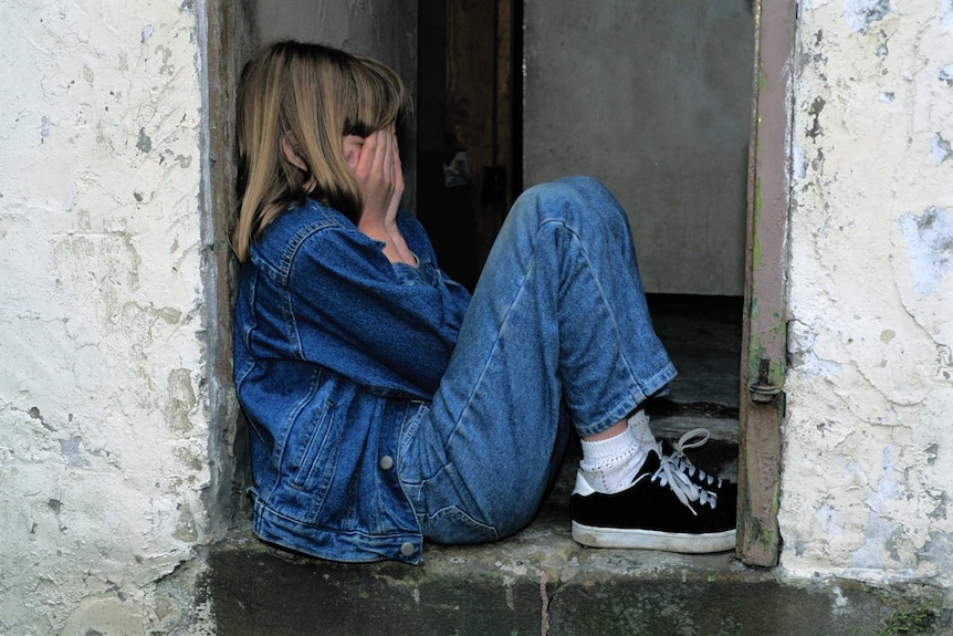 A girl wearing a blue outfit sits in a doorway while covering her face with her hands.
