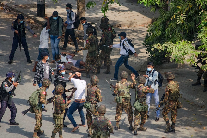 Soldiers and police clash with protesters on a road, where two people are lying down seemingly trying to protect their heads.