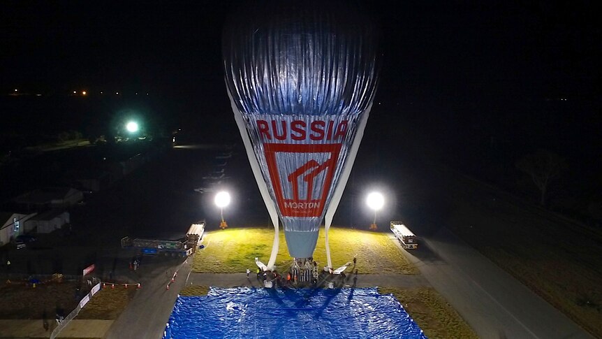 A picture taken from a drone of a tall silver hot air balloon sitting on the ground at night, with floodlights around it.