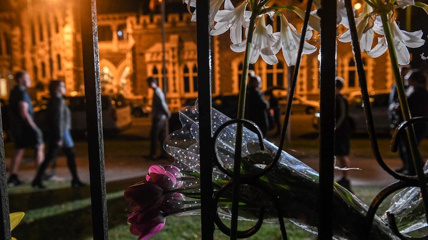 Flowers attached to a fence at night with people walking by in the background.