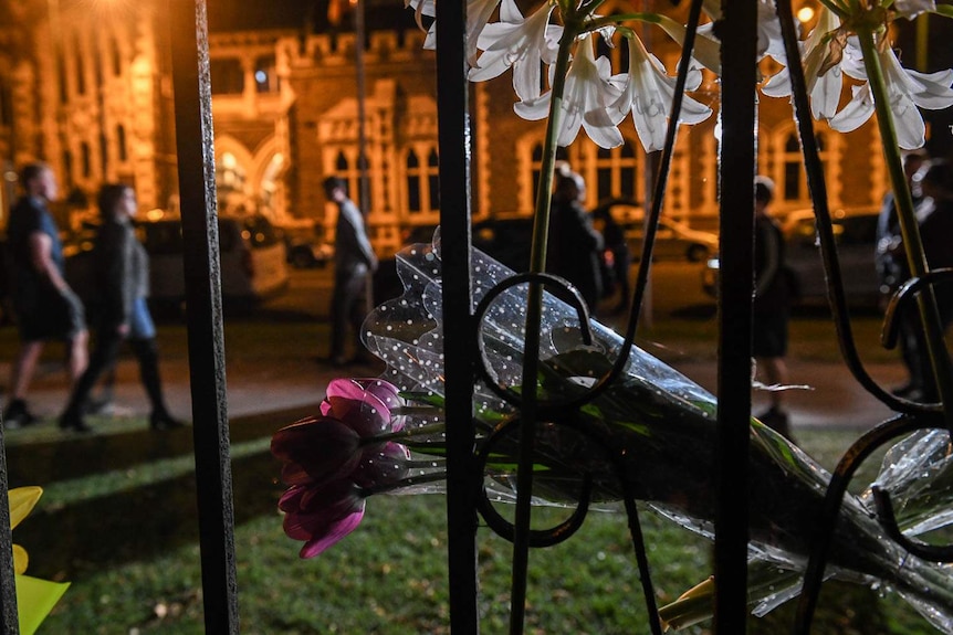 Flowers attached to a fence at night with people walking by in the background.