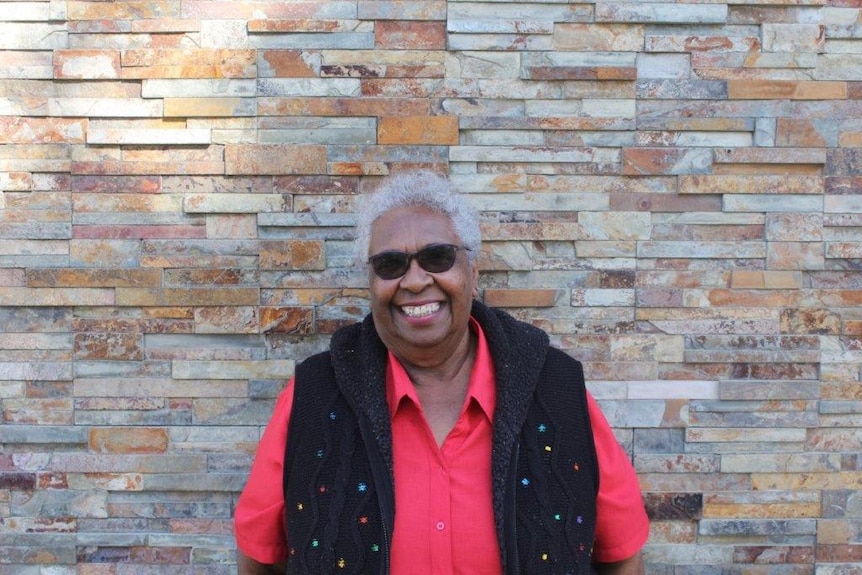 South Sea Islander woman in red shirt and black vest, wearing sunglasses, smiles.