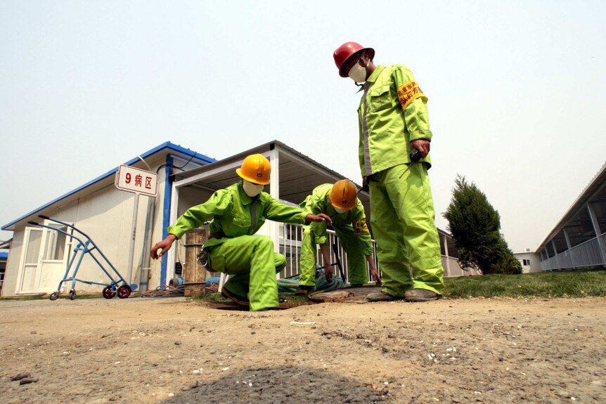Firefighters crouch in yellow suits to check water lines outside a white hospital building.