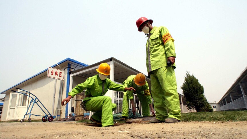 Firefighters crouch in yellow suits to check water lines outside a white hospital building.