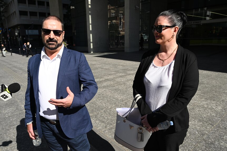 a man and a woman speak into a media microphone outside the brisbane supreme court. both are wearing dark sunglasses
