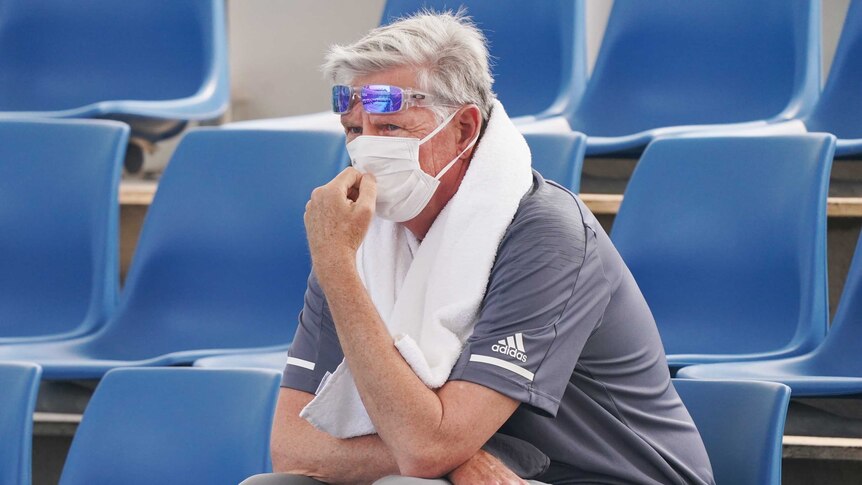 A man sits in the stands and looks on. He has a white face mask over his mouth and nose and a towel around his neck