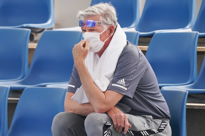 A man sits in the stands and looks on. He has a white face mask over his mouth and nose and a towel around his neck