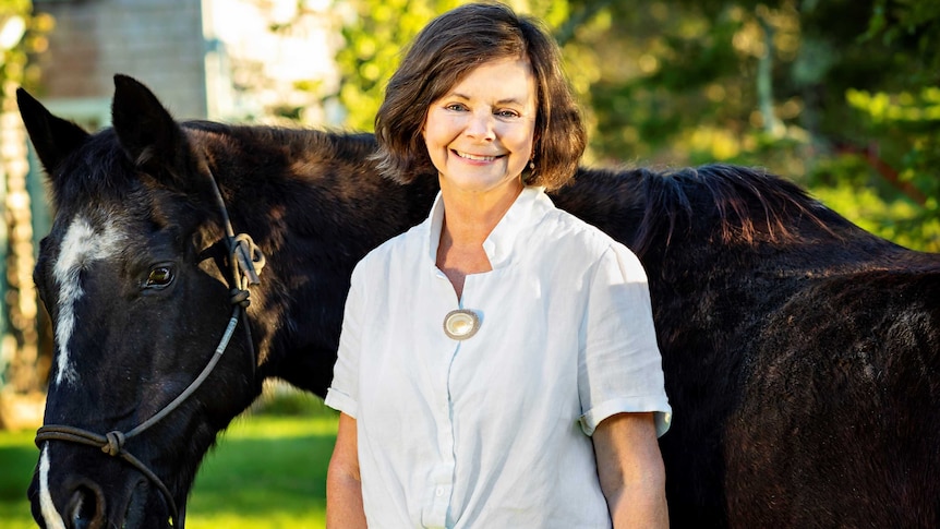 Geraldine Brooks smiling in front of a black horse, trees in the background.