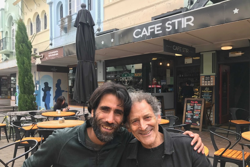 Two men smile, sitting in alfresco dining outside a cafe