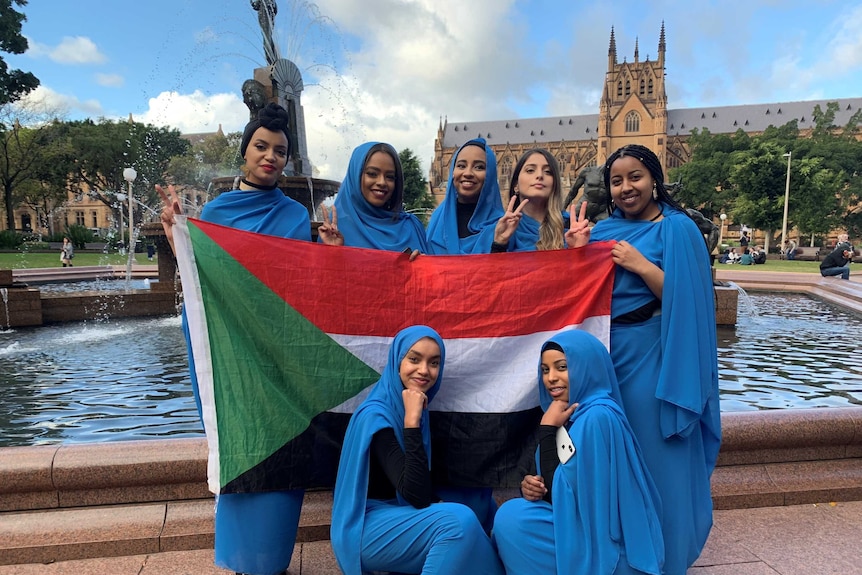 Seven women dressed in blue pose with a Sudanese flag in front of a fountain and cathedral in Sydney