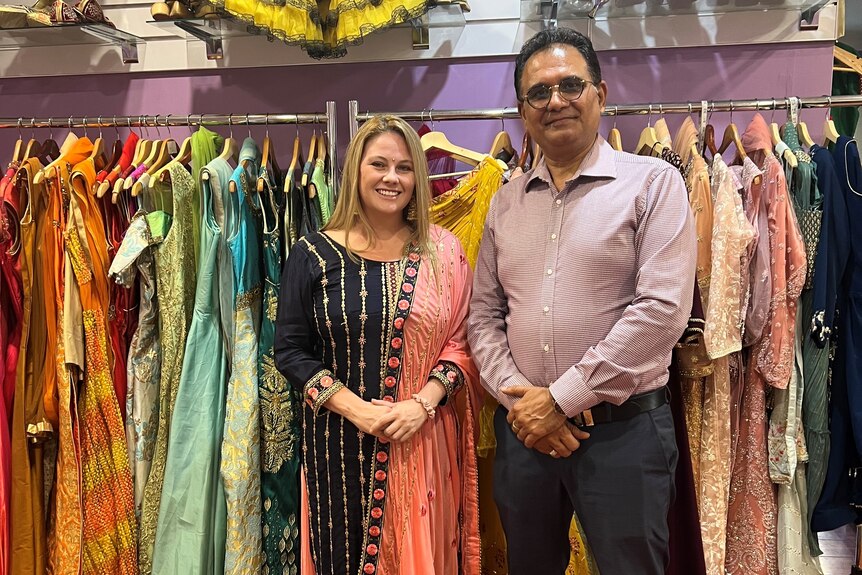 A woman in a sari stands next to a man in a business shirt in front of a rack of saris. 