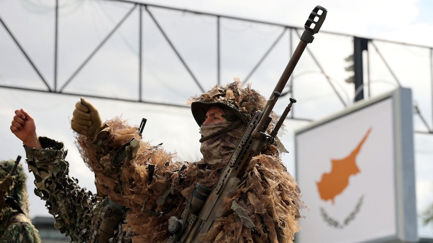 A soldier dressed in camoflague gear holding a gun marches next to an orange sign showing a map of Cyprus