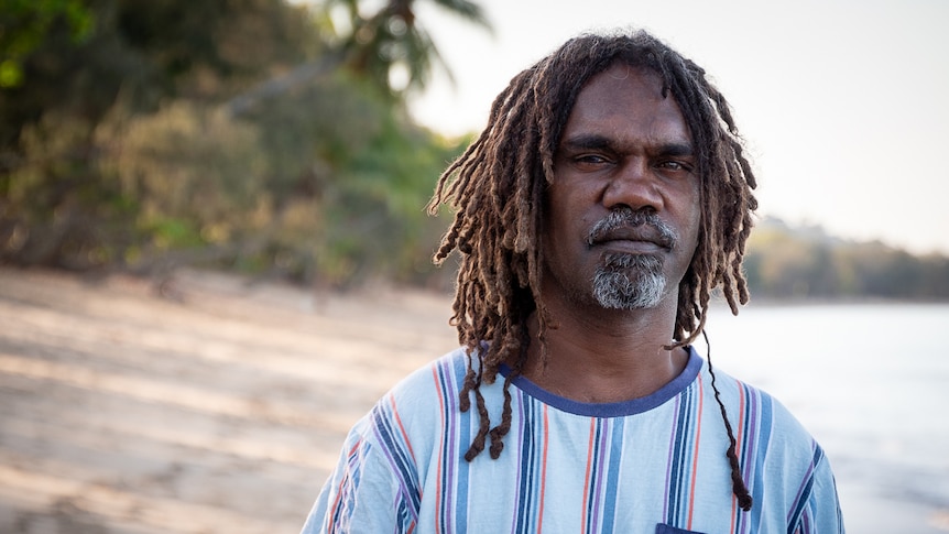 An Indigenous man with dreadlocks and a grey goatee standing on a beach