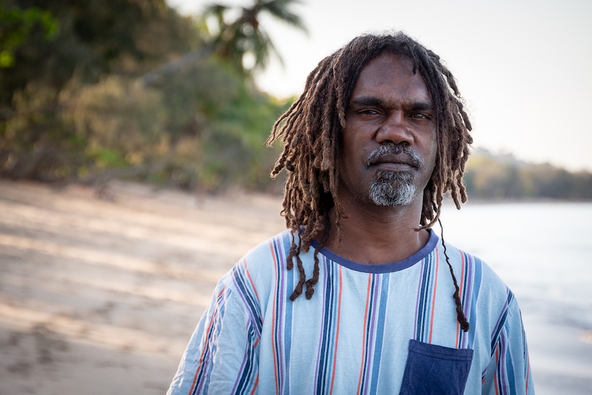 An Indigenous man with dreadlocks and a grey goatee standing on a beach