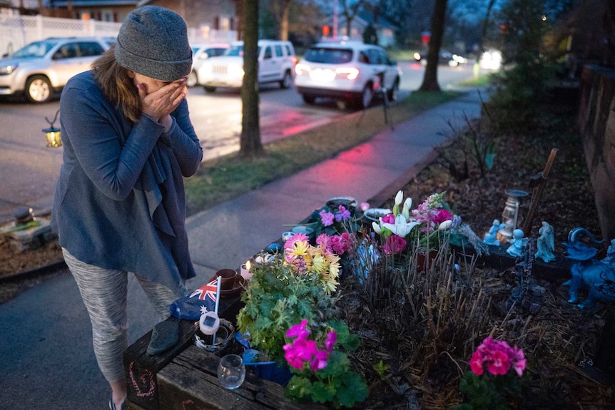 A woman wiping tears in front of a memorial with flowers and a small Australian flag