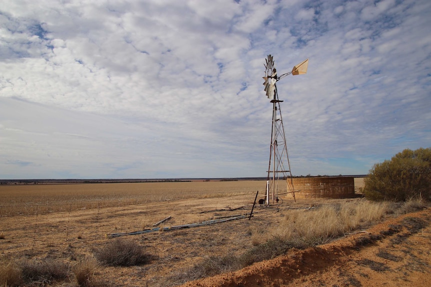 A old windmill stands in a dry paddock on a farm under a cloudy blue and white sky.