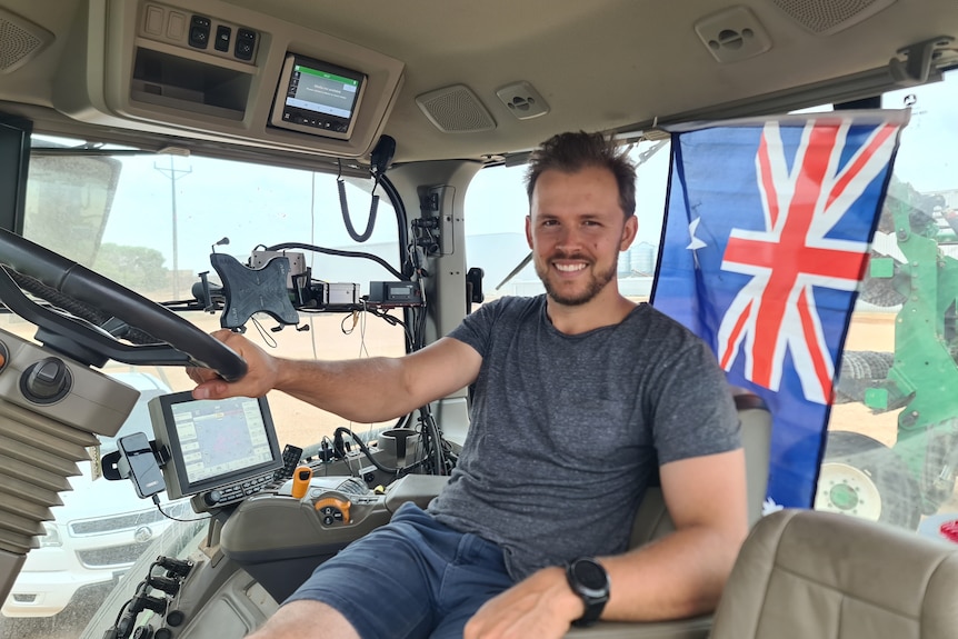 A smiling, dark-haired man in the cabin of his tractor. An Australian flag hangs behind him.