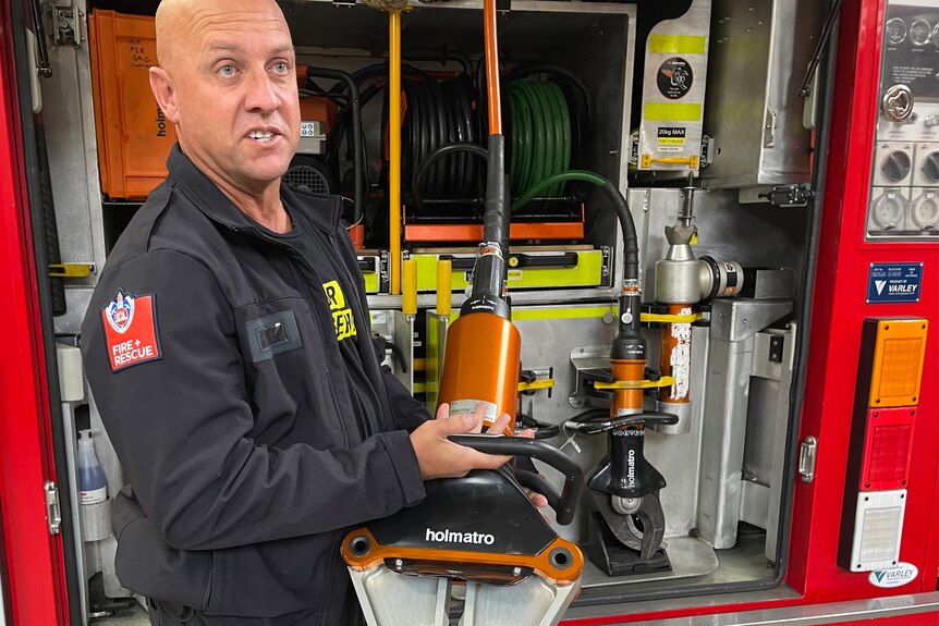 A bald man holding a cutting tool in front of fire truck