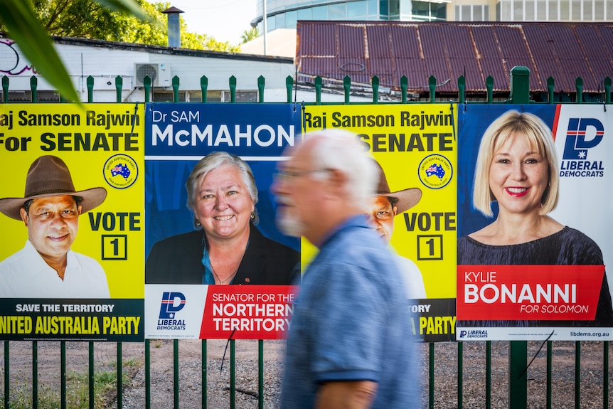 A man walks past a wall of corflutes in the Darwin CBD. One is for Sam McMahon, who is running for the Liberal Democrats.