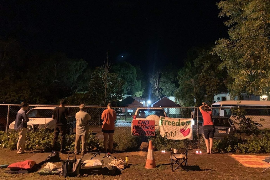 Five refugee advocates stand against a high wire fence with their backs to the camera. Signs read 'end indefinite detention'.