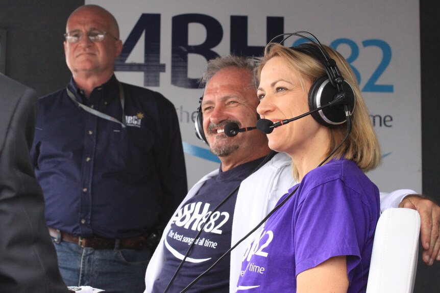 A man and woman wearing radio headsets with handset mics smile in front of a 4BH sign.