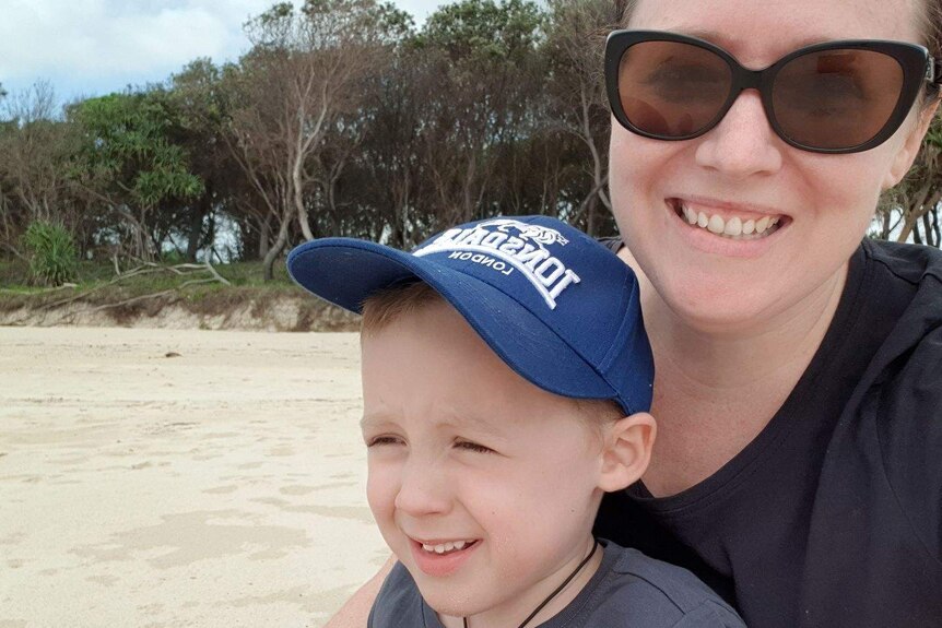 A 30-something woman with sunglasses, holds a small boy in a blue cap on a beach with trees behind.