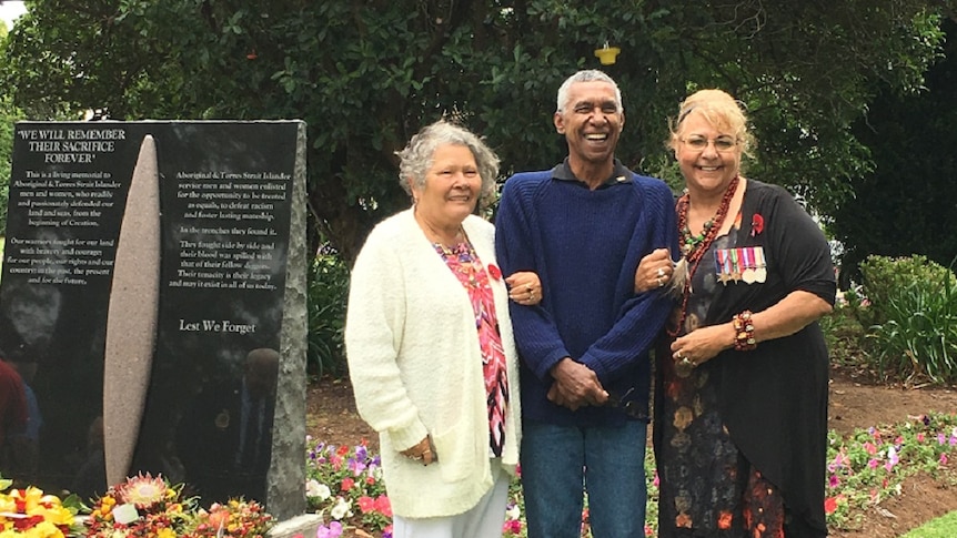 Two Indigenous Elders (women) stand with an Indigenous man in front of a war memorial