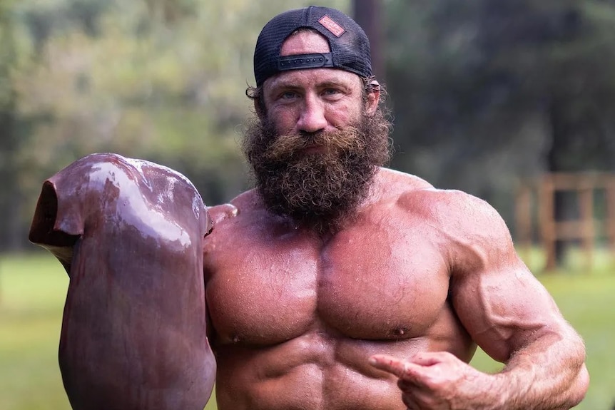 Highly muscular shirtless man points at large raw animal liver while standing outside