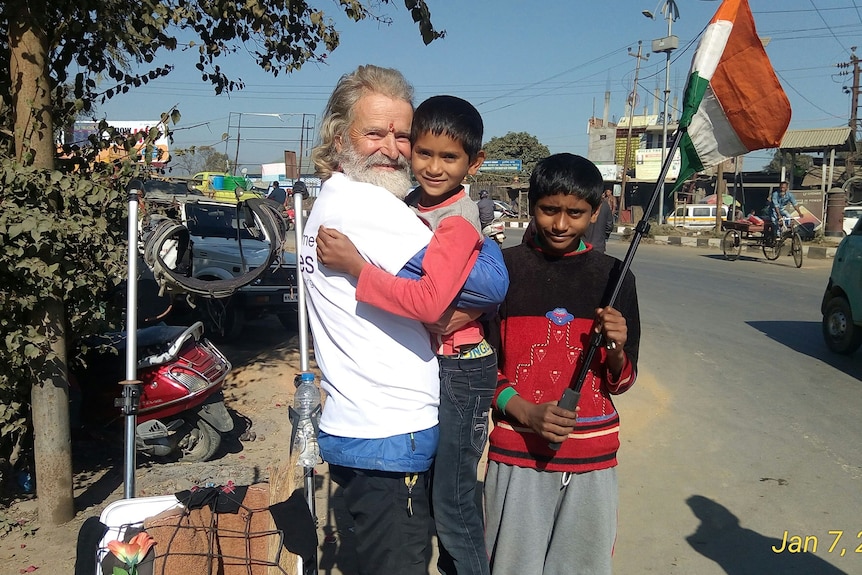 Kuno is holding a young boy on the side of the road, while another boy stands holding an Indian flag.