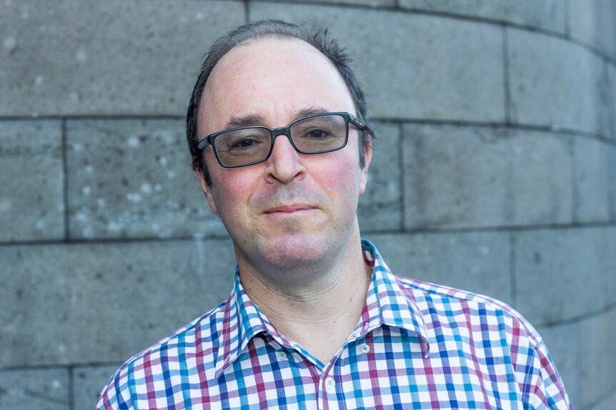 A balding man wearing glasses and a checked shirt looks at the camera, stone wall in background.