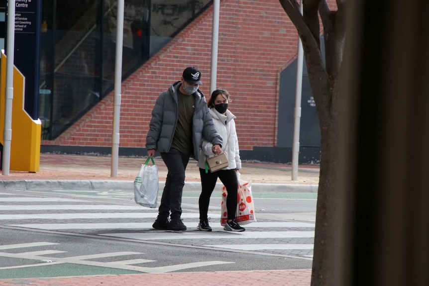 A couple wearing masks hold hands as they cross an empty road.