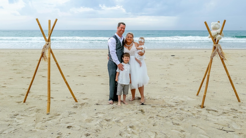 A young man, a boy, and a young woman, holding a toddler, stand on white sand on a beach for a wedding vow renewal ceremony.