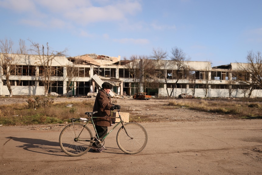 A man rides his bike in front oif a building that is partly destroyed. 