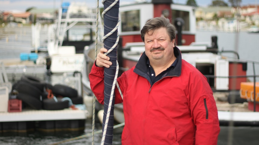 A man with brown hair, wearing a red outdoors jacket, standing next to a mast, with a harbour in the background.