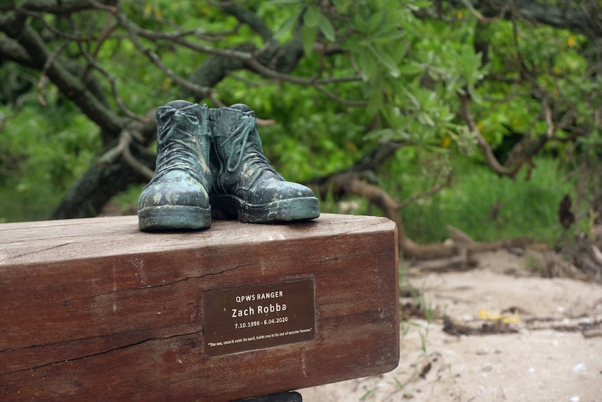 Two brass boots sitting atop a wooden bench, sign says QPWS RANGER, Zach Robba, greenery, sand behind.