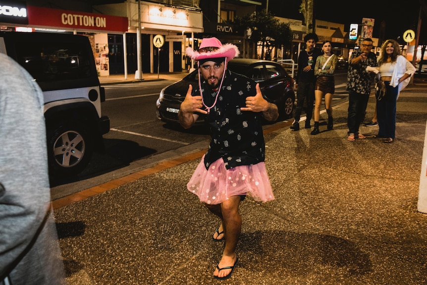 A man gives a thumbs up while wearing a pink cowboy hat, pink skirt and a pair of thongs.