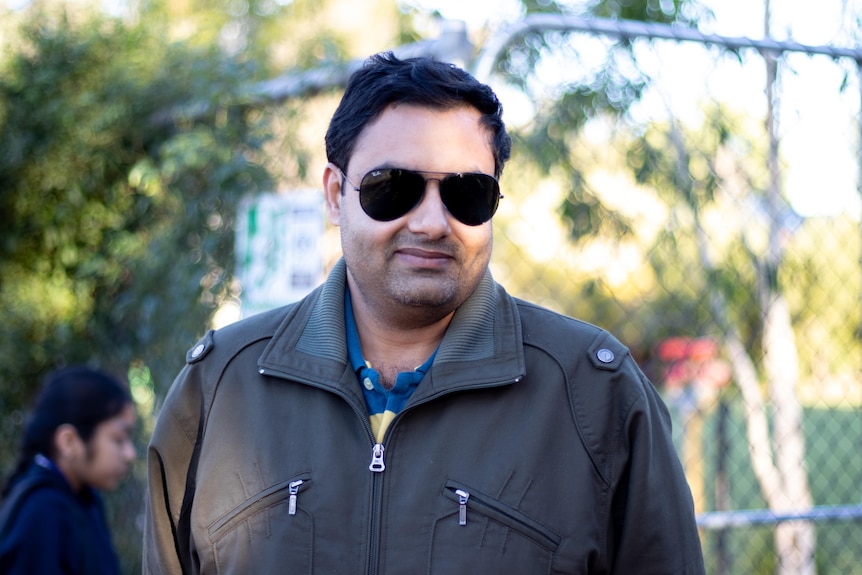 A mid-shot of a man wearing sunglasses and a grey jacket smiling outdoors.