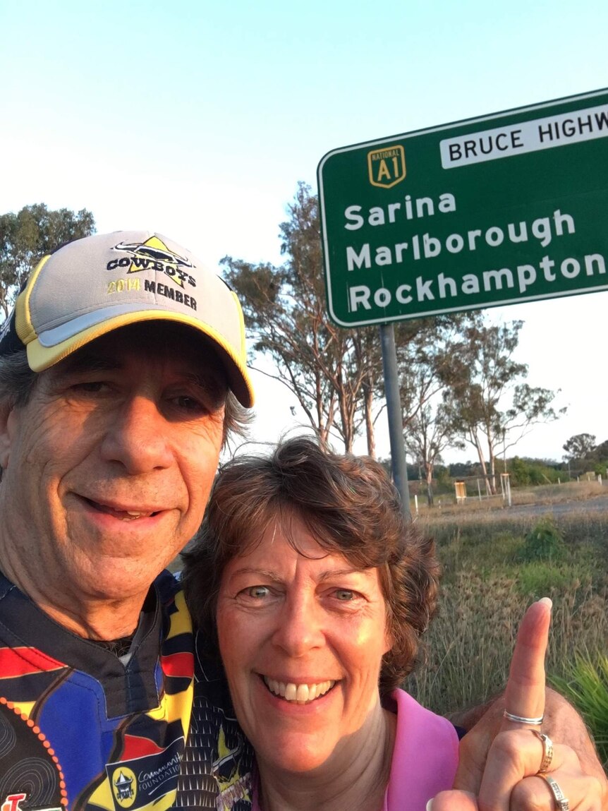 Peter Rowe and his partner Marion smiling and pointing to a sign on the Bruce Highway near Sarina, Queensland.
