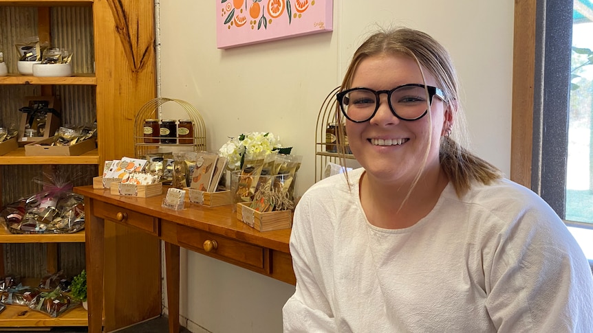 A young blonde woman wearing glasses sits smiling next to furniture bearing her products.