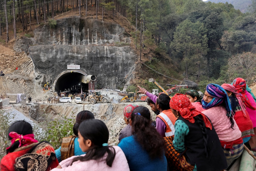 A dozen people wearing colourful clothes sitting on the side of a moutnain looking towards a tunnel opening in a mountain