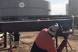 A welder working on a pipe, with a large storage tank in the background
