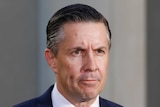 A man with slicked back brown hair wearing a suit and tie with a stern look on his face