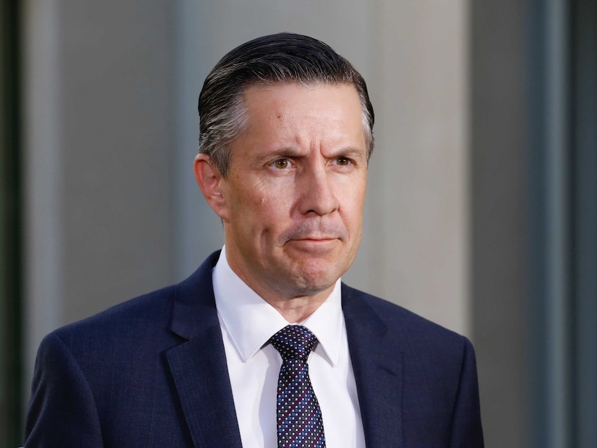 A man with slicked back brown hair wearing a suit and tie with a stern look on his face