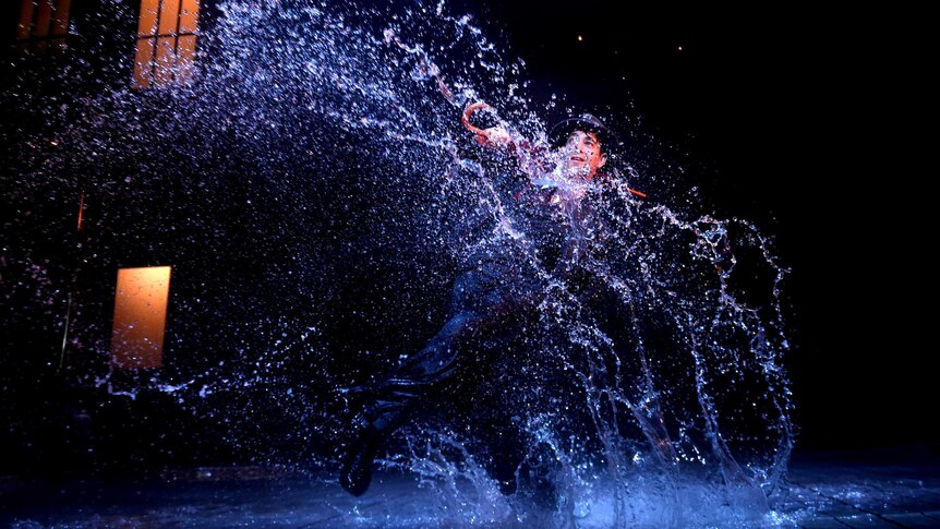 Adam Garcia kicks the water on a darkened stage with a foot, causing it to spray in the air.