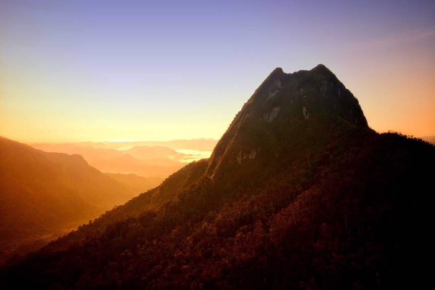 Golden glow at sunrise over sharp peaked mountain with blue sky in distance.