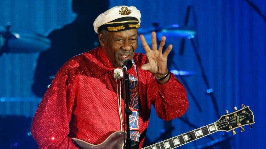Musician Chuck Berry stands on stage in front of a microphone while holding a Gibson guitar