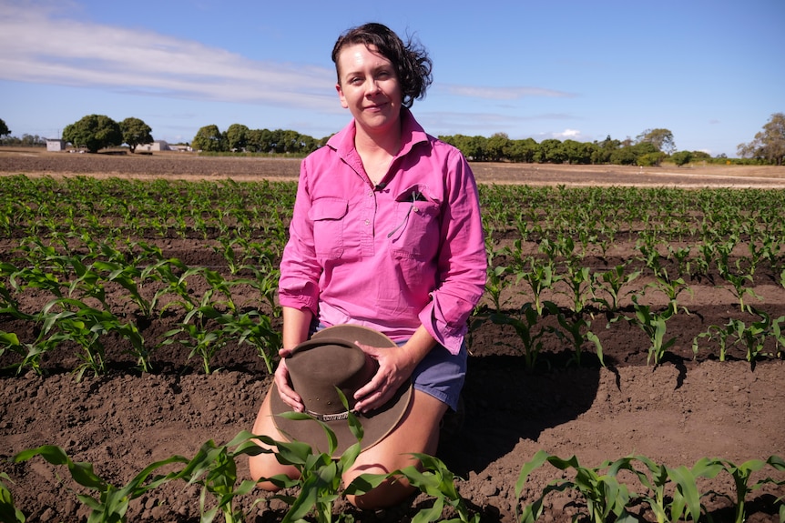Jessica Volker kneels in a field with rows of seedlings sprouting in it, she holds her hat in her hands