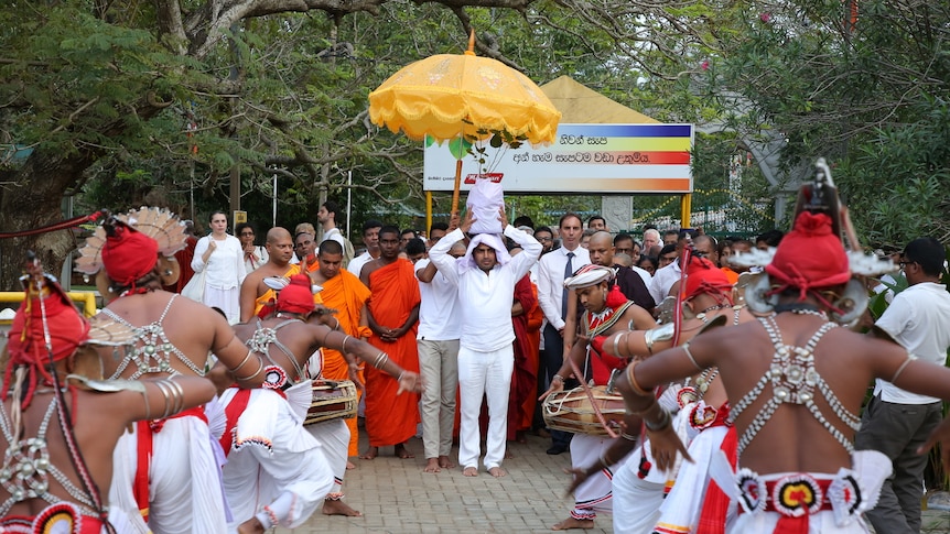 A colourful ceremony with people farewelling the sapling that is being held on a man's head. It's protected by a parasol