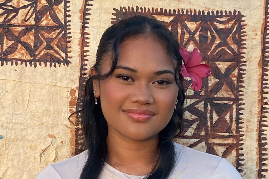Young Samoan woman wears pink flower behind her left hair, white tee, standing infront of tapap cloth. 