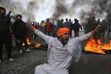 A protestor shouts slogans as fire burns and smoke rises in the background.